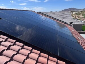 All-black solar panels on a red clay tile roof of an Arizona home, with neighboring tile roofs, desert landscaping and rugged mountains under a blue sky