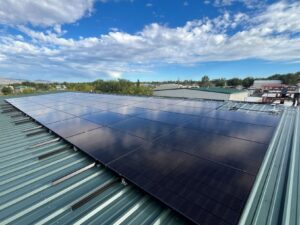 Rows of black solar panels mounted on a green metal roof under a blue sky with scattered clouds, with mountains on the horizon in Arizona