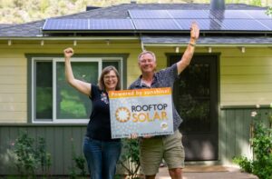 Smiling couple cheering and holding a Rooftop Solar “Powered by sunshine!” sign in front of their yellow Arizona home with solar panels on the roof